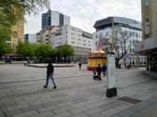Foto des Meidlinger Platzes. Einige Menschen sind zu sehen (tw. mit Kinderwagen). In der Mitte der Brunnen, rechts ein geschlossenes Karussell. Im Vordergrund ein Umgebungsplan.